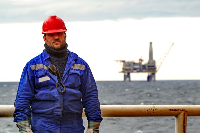 A person in a blue work suit with an oil rig in the background.