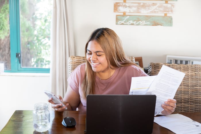 Smiling young person sits at a table in front of laptop and papers while using cell phone.