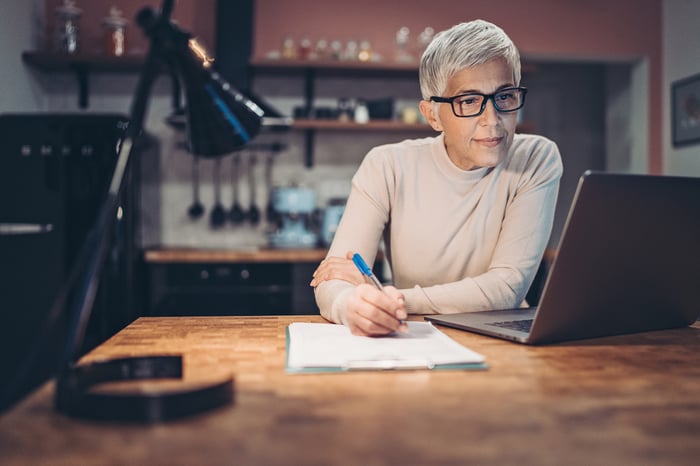 Mature adult sits at a desk in front of a desk lamp and laptop.