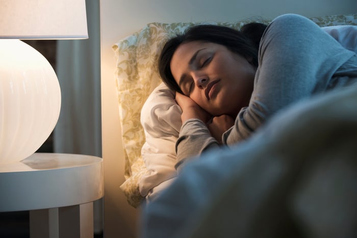 A woman sleeping on a bed with a lit lamp on a night table.