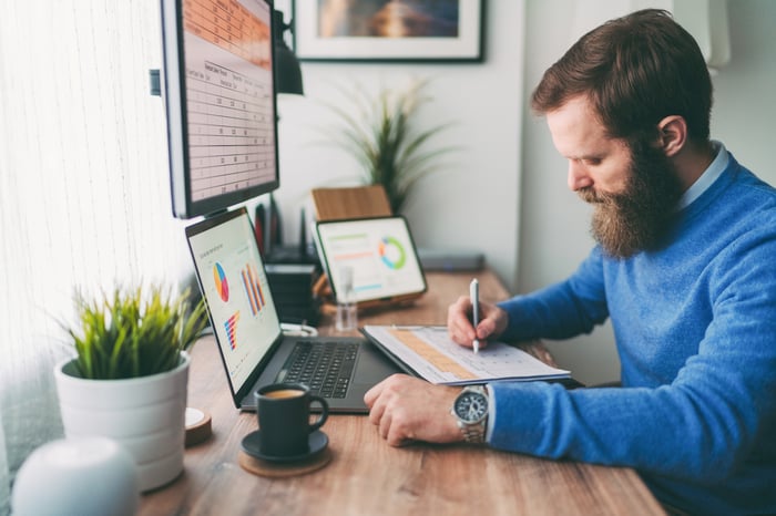 Adult works at a desk in front of a laptop, monitor, and cup of coffee.