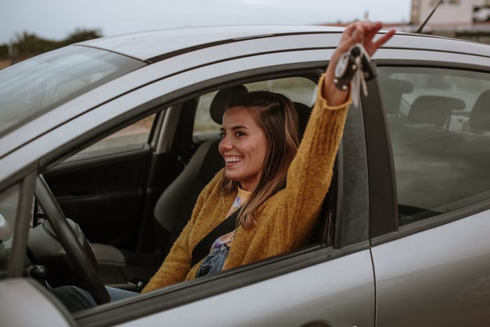 A smiling young woman sitting in her car, holding the keys out the window.