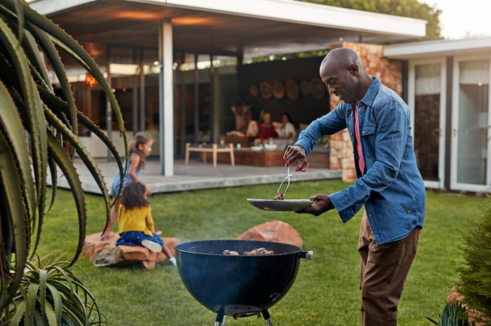 A person grilling in the backyard while family members play in the yard and sit under a patio.