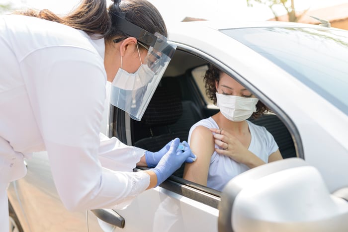 A masked, gloved, and shielded person administers a shot to a masked person in a car.