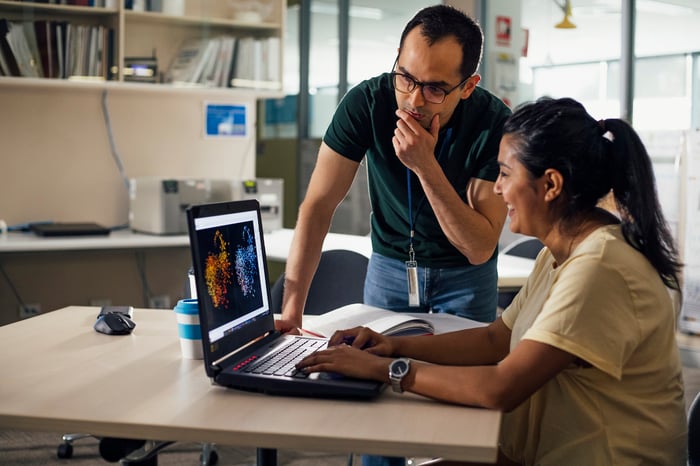 Two people in an office setting look at a laptop screen.