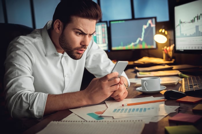 A confused person looking at his phone with computer screens displaying stock price charts in the background.