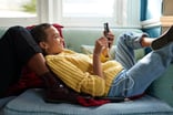 Young person loungs on another person's legs on a blue couch while scrolling through their phone.