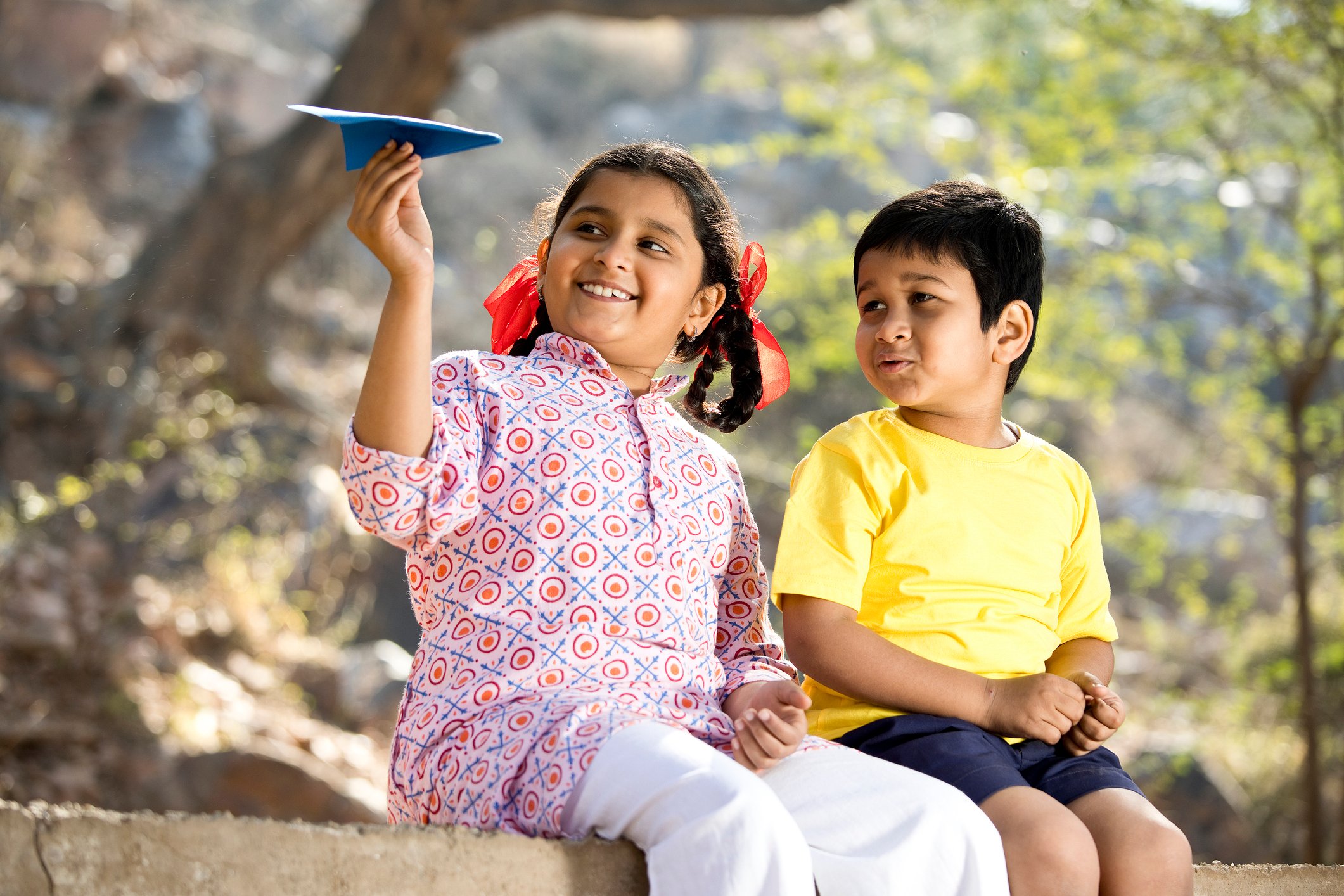 A child holding a paper air plane sitting next to another child.