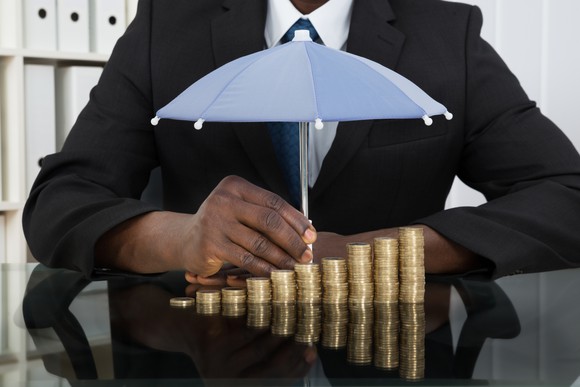 Person in a suit holding a small umbrella over a stack of coins.