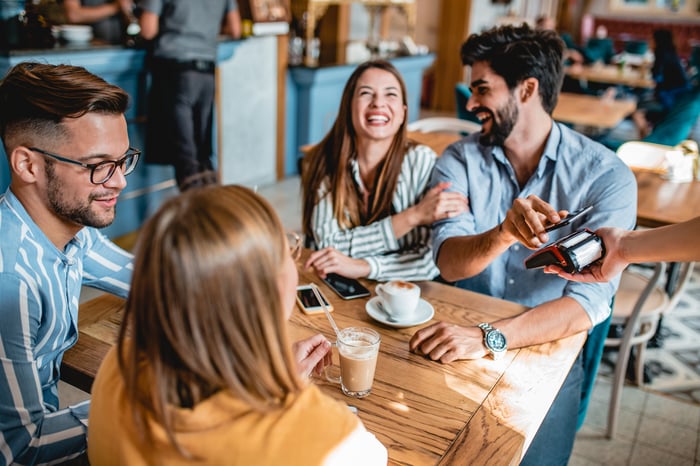 A group of people at a restaurant and one paying with a mobile phone.