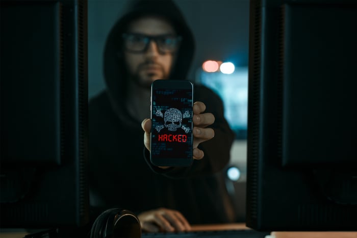 Hooded person holding a phone that says "hacked" on the screen.