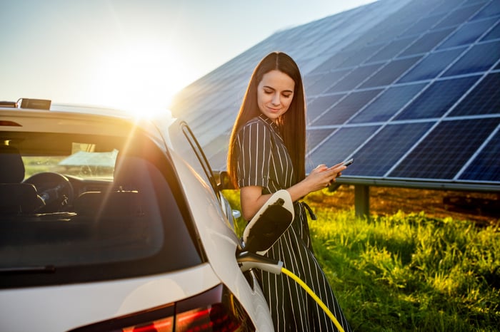 An electric-vehicle owner with solar panels in the background charges a vehicle. 