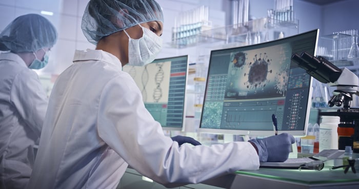 Masked lab worker takes notes while looking at virus on computer screen.