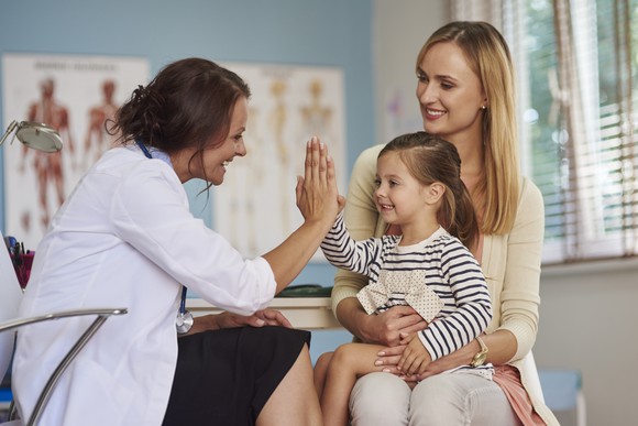 A physician giving a high-five to a child seated on her mother's lap.