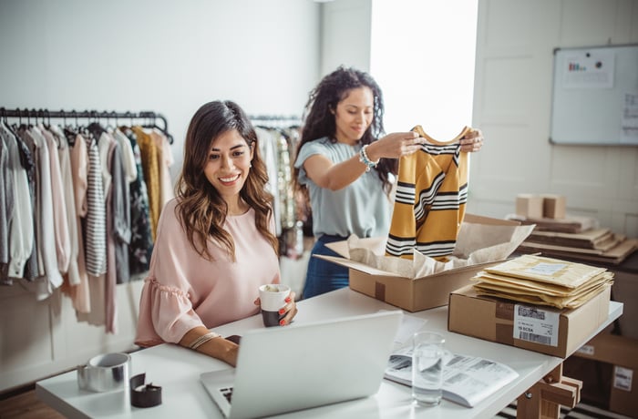 Two people in a shop, one using a computer, and the other opening a garment box.