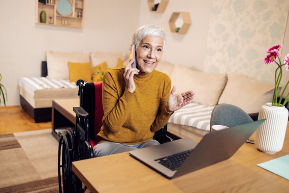 A woman sitting in a wheelchair at a desk with a computer and talking on the phone.