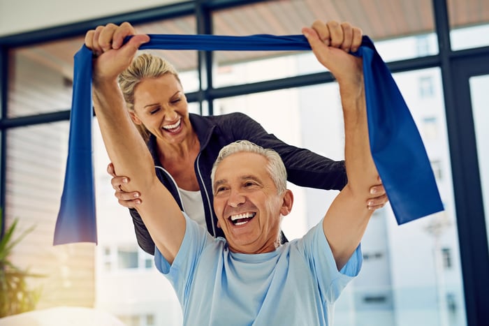 Older patient smiling as he works with a strength band with help of smiling physical therapist/