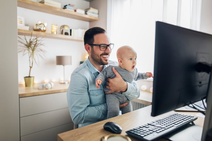 Smiling person holding baby at desk with computer.