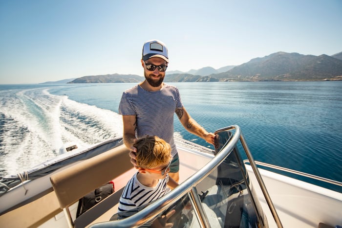 Adult on a boat with a child steering it through a large body of water with mountains in the background.