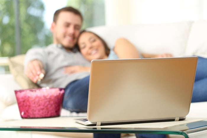 A couple laying on the couch eating popcorn and looking at a laptop.