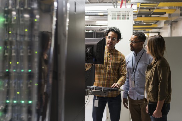 Three people work on a computer in a data center.