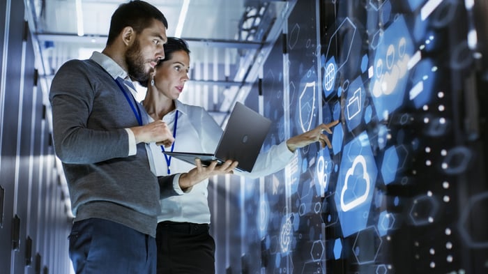 Two people standing in a data center with a laptop, analyzing a digital screen.