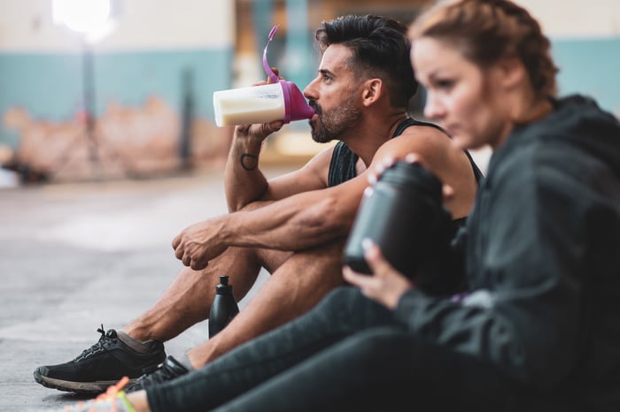 Two people in a gym drinking protein shakes.