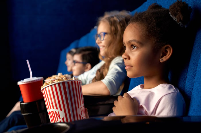 Young children eating popcorn and holding drinks in a movie theater. 