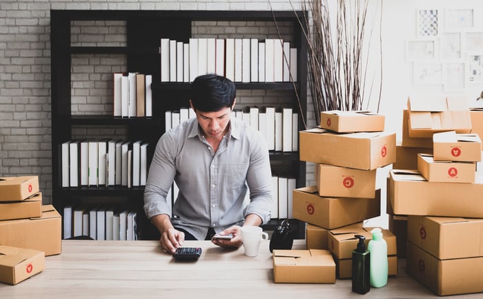 Person surrounded by shipping boxes crunching the numbers for their small business. A black bookcase and grey brick wall are in the background. 