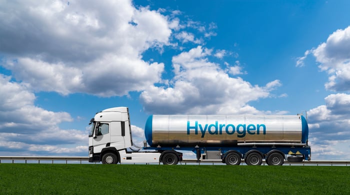 Tanker truck labeled Hydrogen drives along a road under a blue sky with clouds.