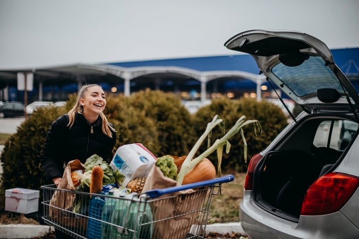 A person with a shopping cart filled with groceries standing next to the open hatchback of a car.