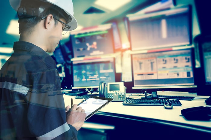 An oil industry worker sitting at a desk with a hard hat on, working on computers.