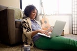 Young adult browses on a laptop while sitting on the floor beside a guitar and mug.