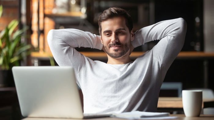 A relaxed man with his hands behind his head slightly smiling at a laptop.