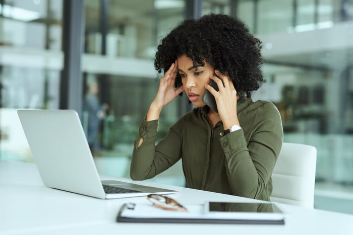 A concerned woman talks on a smartphone while looking at a laptop.