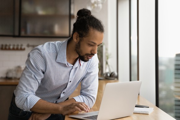 An investor looks at something on a laptop on a counter near windows.