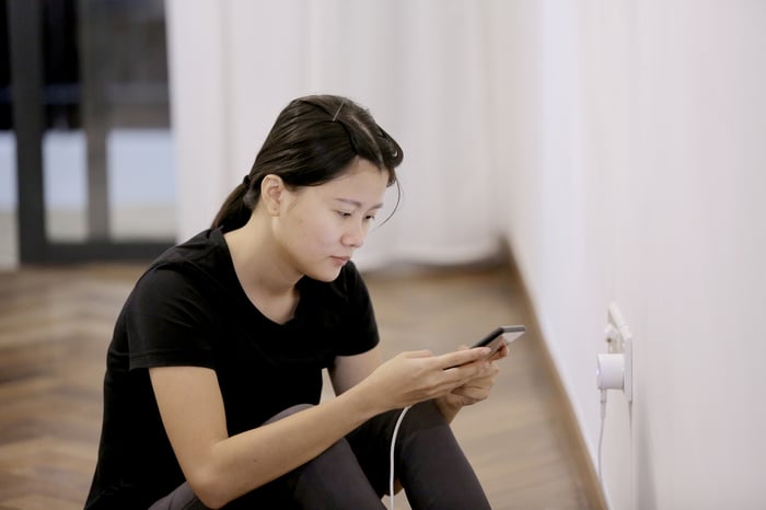 A person sitting on the floor looking at a smartphone while it's charging.