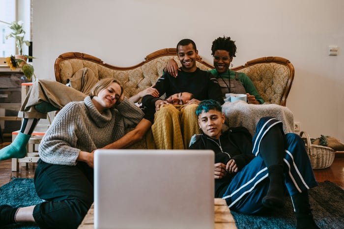 Five young adults sitting lounging about a living room watching streaming on a laptop.
