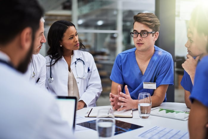 A doctor talks with several of his colleagues as they sit at a table covered in binders and papers.