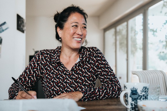 A lady smiling while sitting at a wooden desk with a pen in her right hand.