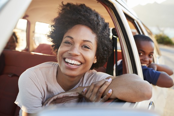 A smiling parent leaning out the driver's side window of their parked car, with their kids in the back seat.