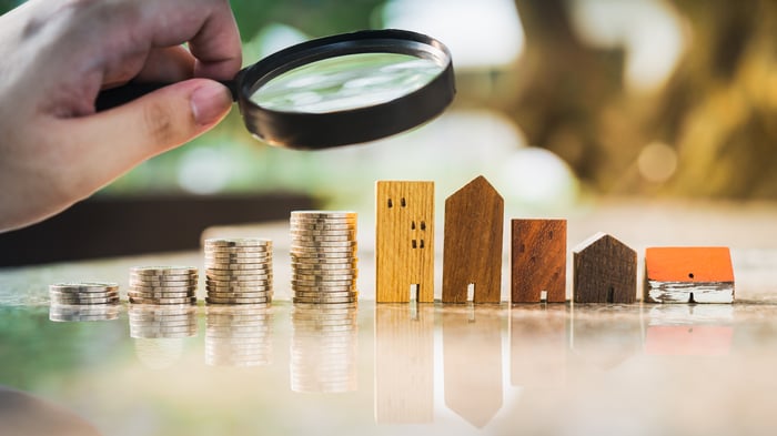 A person holding a magnifying glass looking at a row of miniature buildings next to a set of rising coins. 