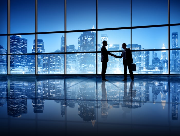 Two people shake hands with an office skyline in the background.