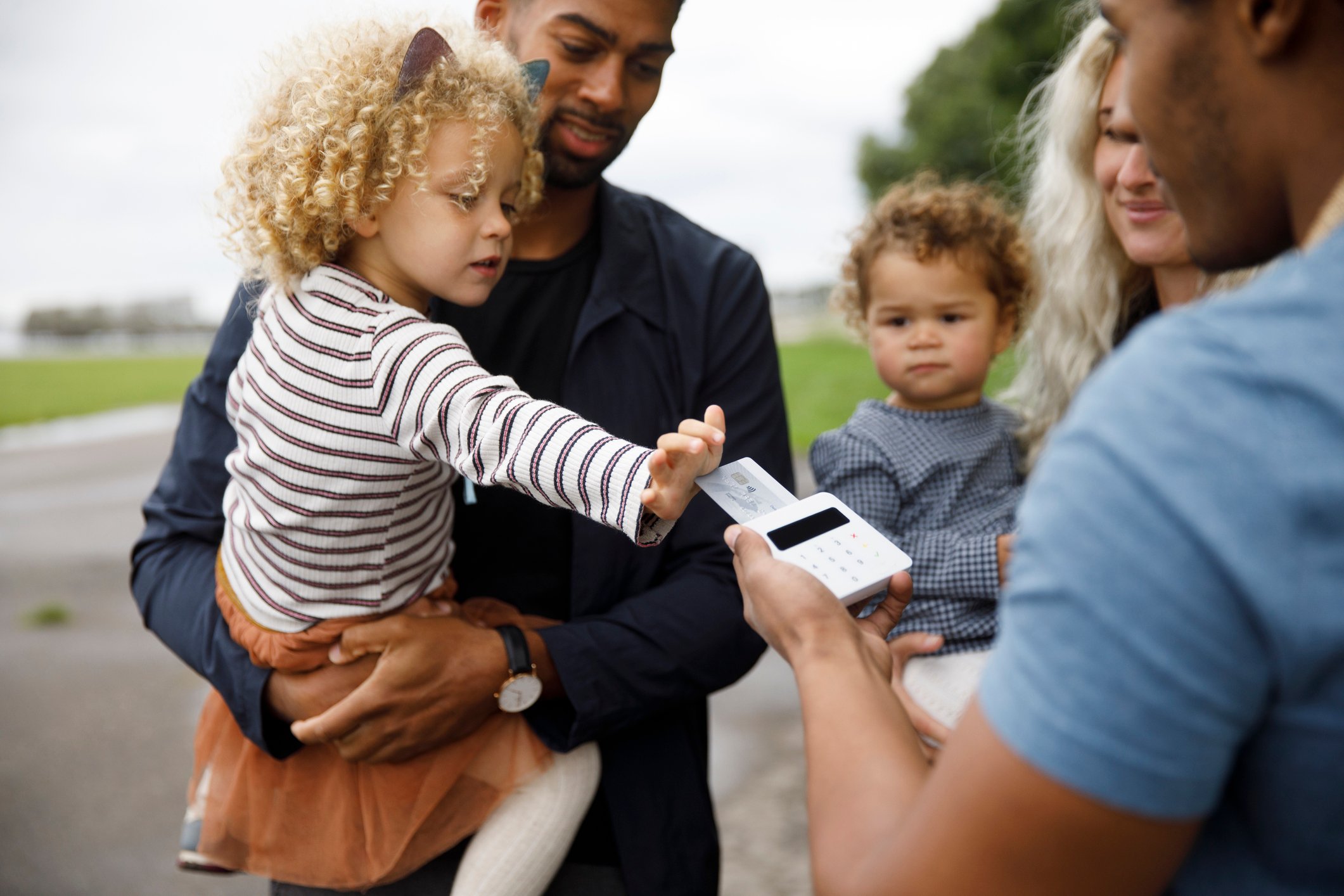 A family standing outside with a father holding a child who is using a credit card in a terminal that a person is holding