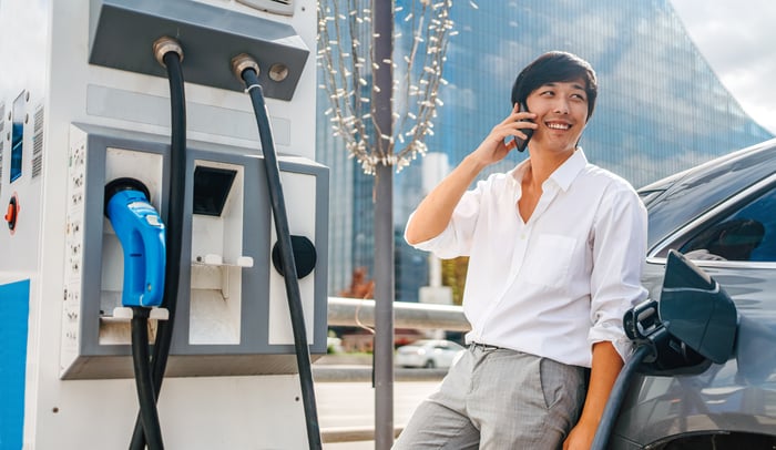 Young person charging an electric car at a public charging station while talking on the phone.
