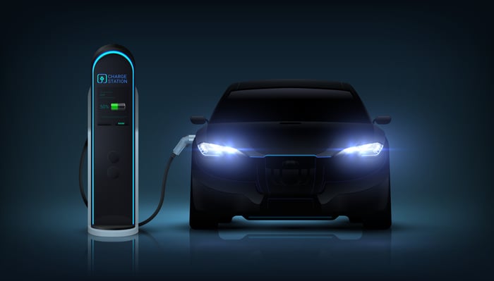 With headlights glowing, an electric car  is plugged into a charging station.