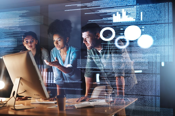 A team of employees huddle around a computer, analyzing and discussing data on the screen.