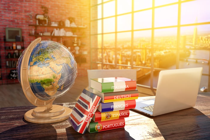 Language dictionaries stacked on a desk between a globe and a laptop, in a warmly lit room overlooking a city.