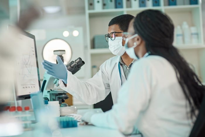 Two people seated at a lab desk featuring a PC screen and microscope.