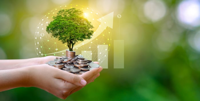 A person holding a pile of coins with a tree growing on top behind a rising chart.